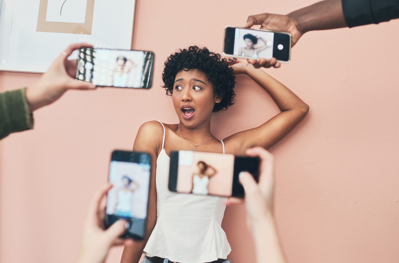 A young woman posing playfully in front of a pink wall while surrounded by multiple smartphones filming her, representing influencer culture and emerging social media trends in content creation.